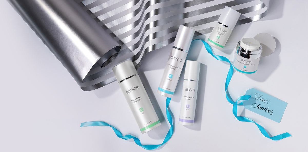 A set of five sanitas skincare products  arranged on a light gray surface with a metallic silver gift wrapping paper, a blue ribbon, and a small gift tag that reads Love, Sanitas.