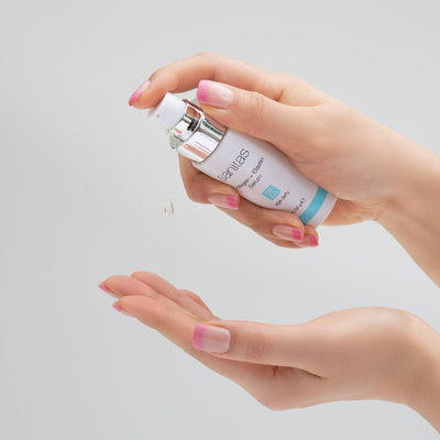 Hand holding Collagen + Elastin Serum with another hand underneath with drop of serum midway on a light gray background