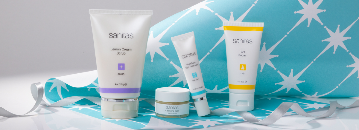 A set of four Sanitas skincare products arranged on a light gray surface with a blue and white star-patterned wrapping paper behind them and a silver ribbon