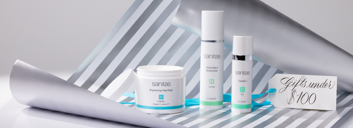 Sanitas skincare products on white surface with diagonal gray & white striped wrapping paper with a gift tag that reads 'Gifts under $100' with a blue ribbon