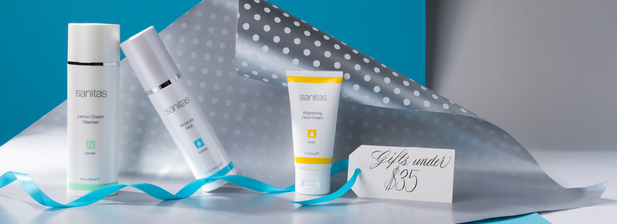 Three Sanitas skincare products arranged on silver gift wrapping paper with white polka dots, blue ribbon, and a small gift tag reading 'Gifts under $35' against a blue and gray background.